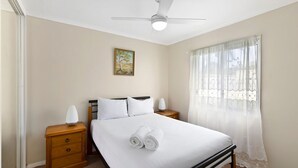 Quarto com cama, mesa de cabeceira, abajur, ventoinha no teto, quadro na parede e janela com cortinas leves.