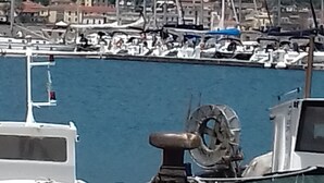 Un port de plaisance avec des bateaux amarrés, une grande ancre au premier plan et une colline avec des bâtiments en arrière-plan.
