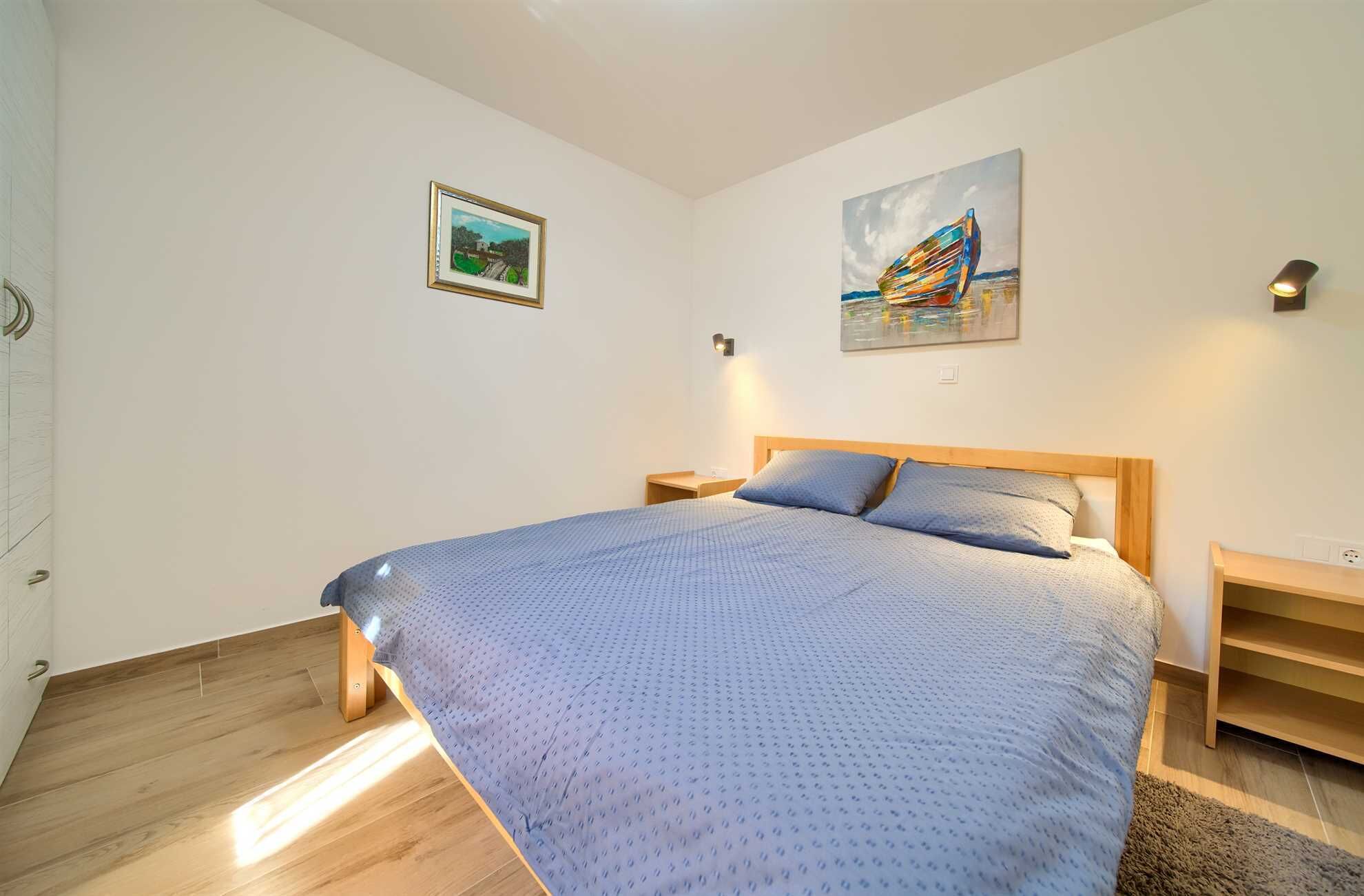 A bedroom with a large bed, wooden headboard, two bedside tables, and two wall-mounted lamps. There are two framed artworks on the walls.