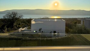A modern building with a flat roof, surrounded by greenery and a view of the sea and mountains in the background.