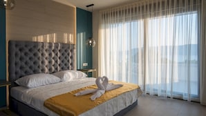 A modern hotel room with a tufted headboard, a large bed with white linens, a gold accent throw, and a view through sheer curtains.