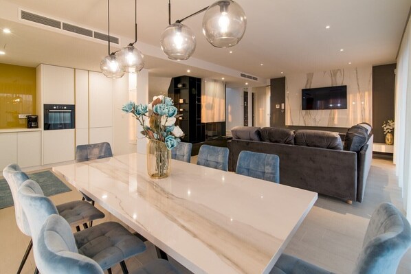 Una sala de estar moderna con una mesa de comedor de mármol, sillas tapizadas en azul, un sofá gris y un florero con flores.