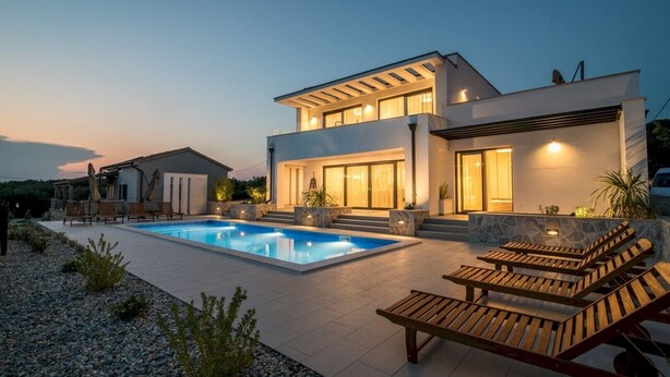 A modern two-story house with a swimming pool, outdoor seating, and a well-lit entrance.