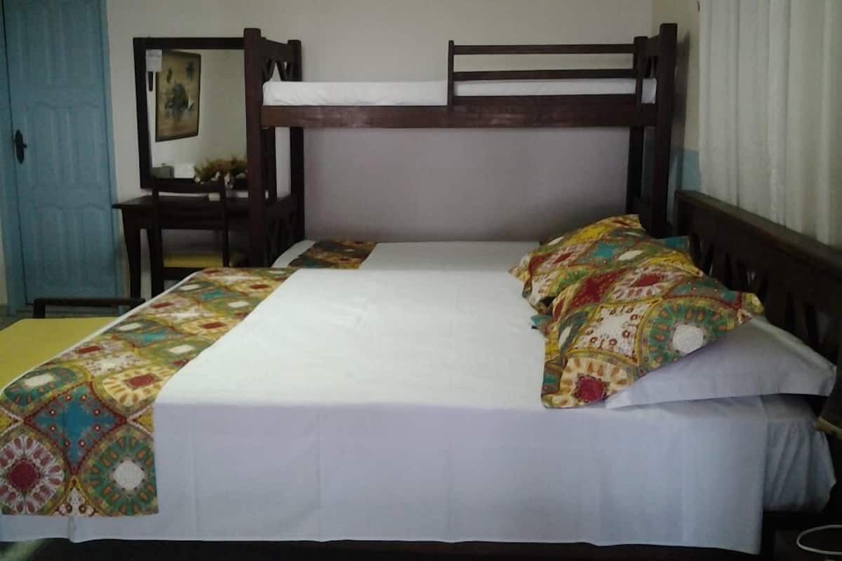A single bed with a wooden frame, a white mattress, and colorful bedding. There is a small table with a chair to the left, and a mirror on the wall.
