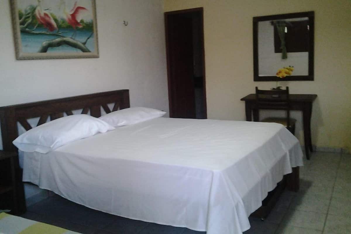 A single bed with white bedding in a room with a wooden headboard. There is a small table with a mirror and a flower vase on the wall.