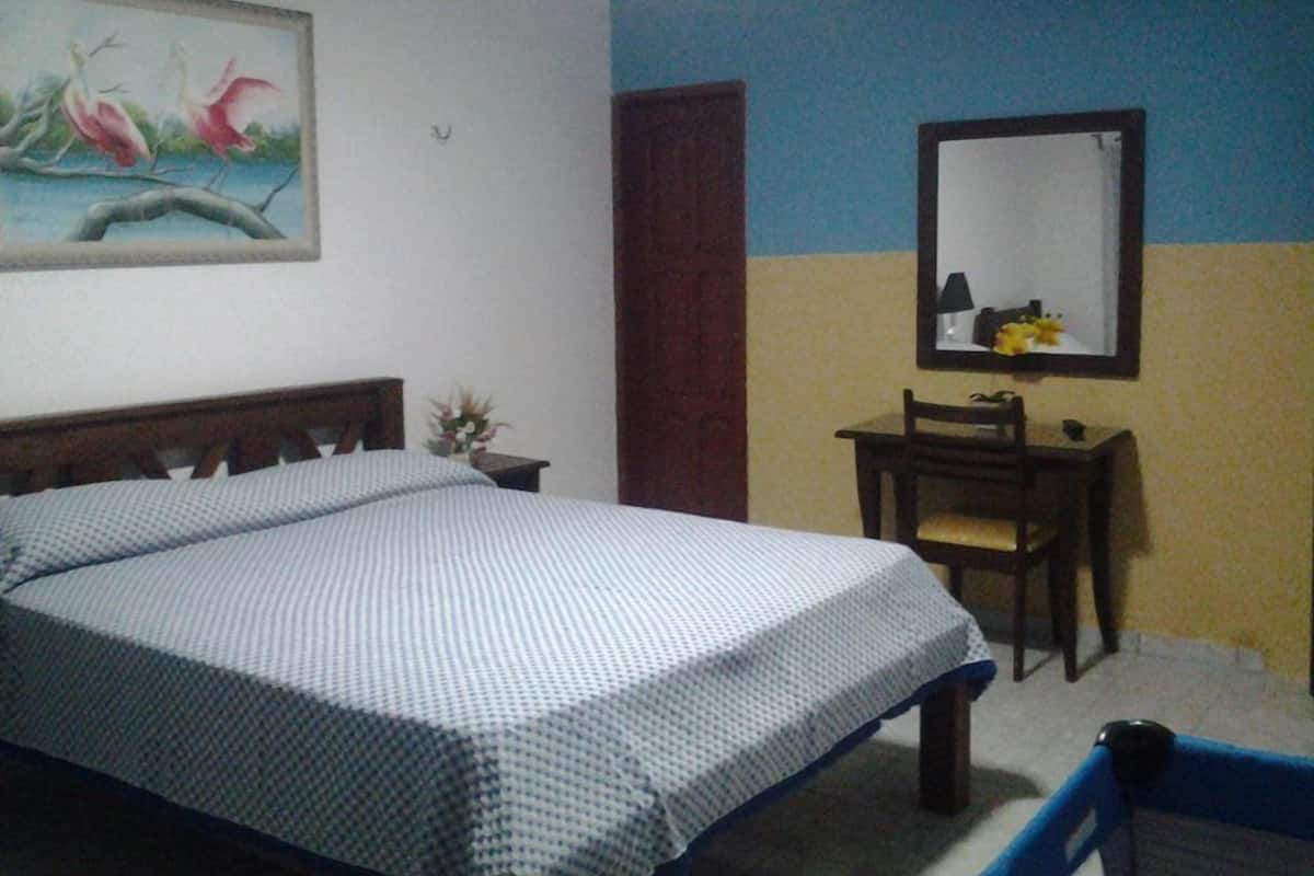 A bedroom with a bed, a desk, a chair, a mirror, and a painting on the wall.