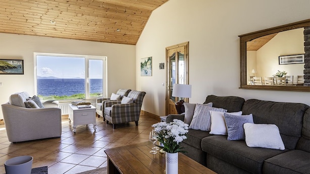 Amplia sala de estar con techo de madera, ventanas grandes con vista al mar y una zona de estar acogedora con un sofá oscuro y dos sillones.