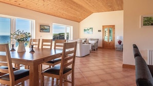 Una habitación amplia con techo de madera, grandes ventanales con vistas al mar, un comedor con mesa y sillas de madera, y una zona de estar acogedora con sofá y sillones.