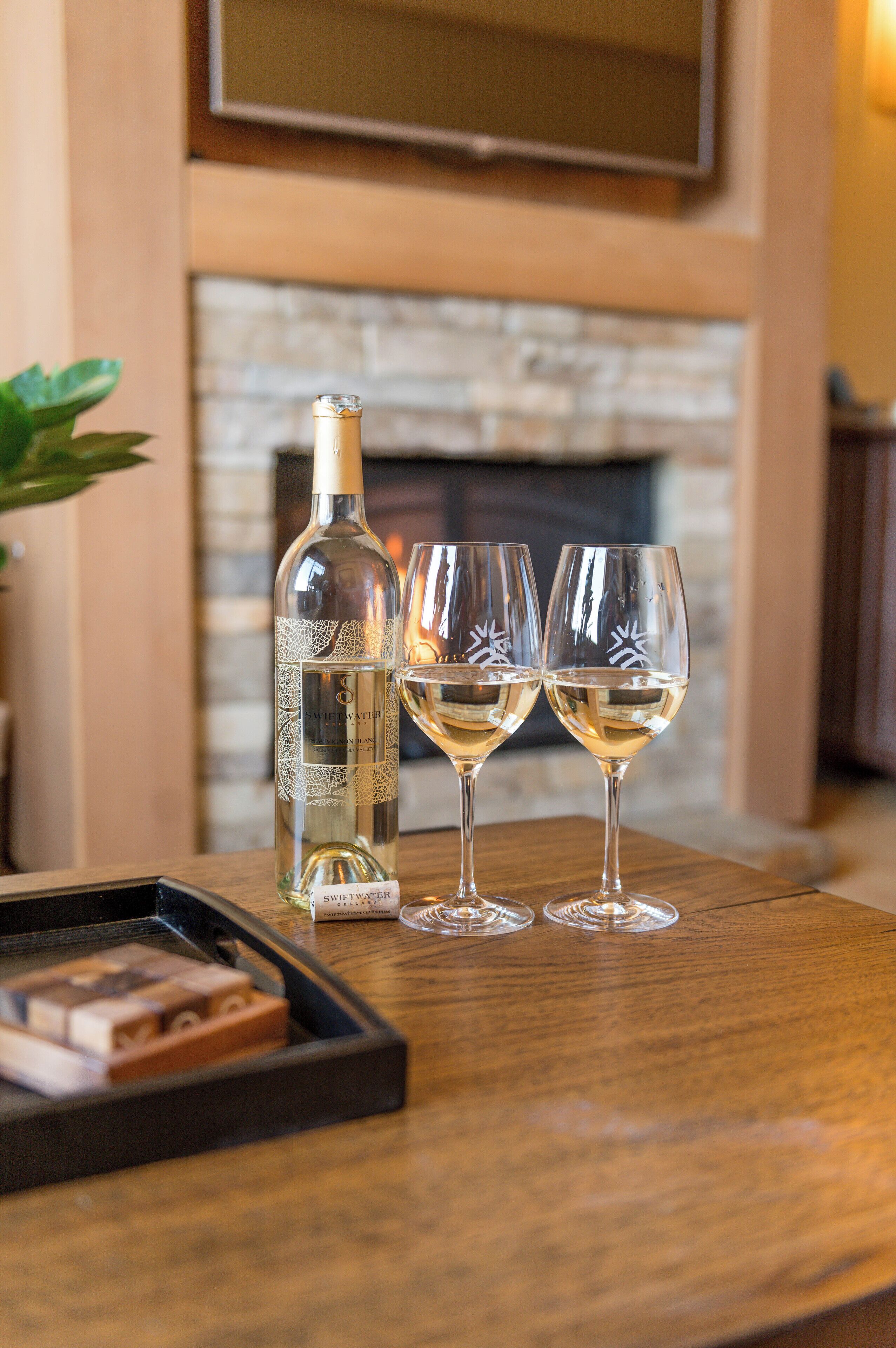 A bottle of white wine and two glasses on a wooden table with a fireplace in the background.