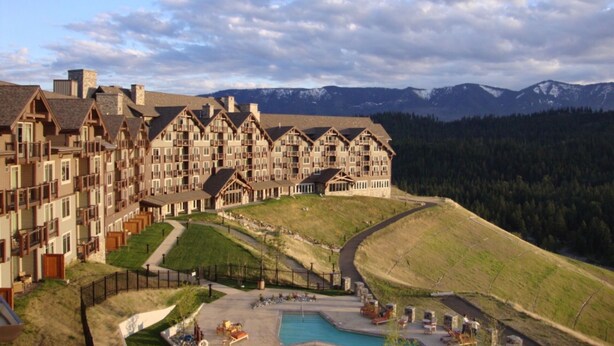 A large resort with multiple chalet-style buildings, a swimming pool, and a mountainous backdrop.