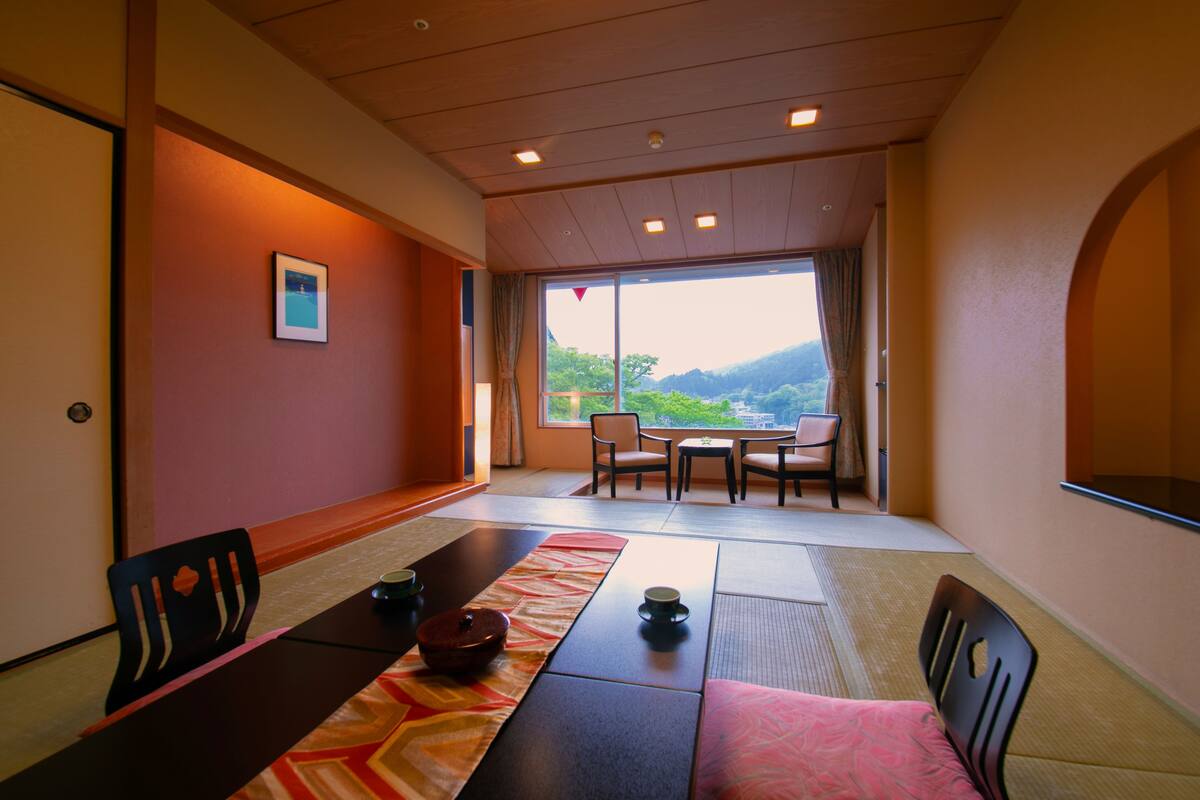 Une pièce de style japonais traditionnel, avec un sol recouvert de tatamis, une petite table sur laquelle est disposé un service à thé, et une vue sur les montagnes, visible à travers les grandes portes coulissantes.