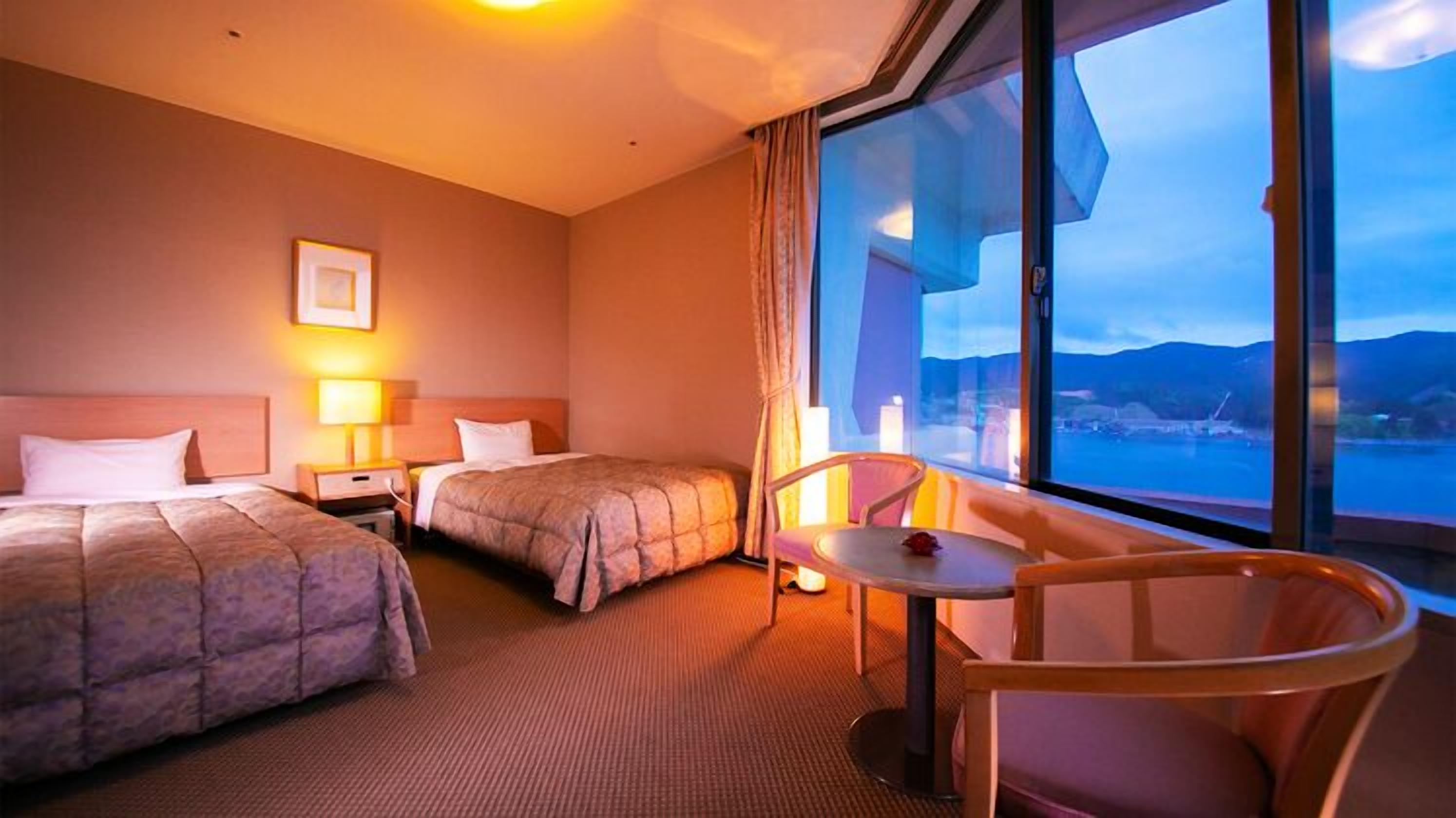 Habitación de hotel con dos camas, una mesita, una silla y un ventanal con vistas.