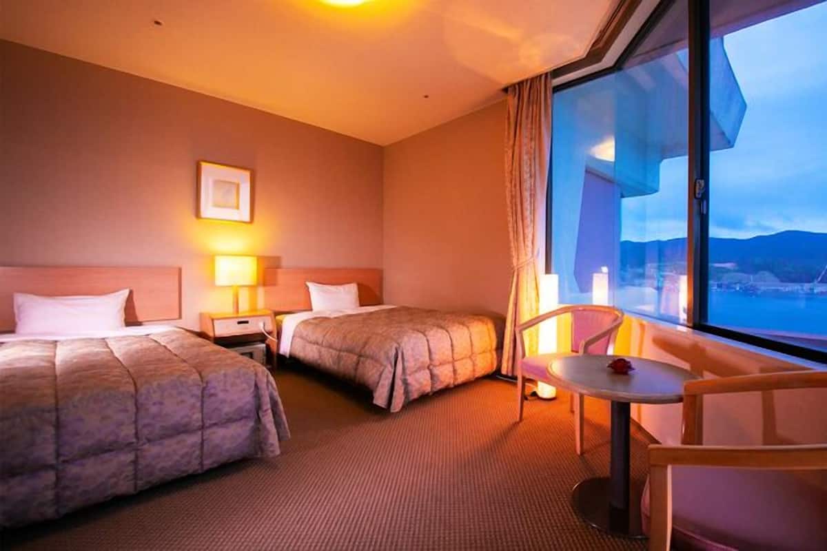 A hotel room with two beds, a small table, a chair, and a large window with a view.