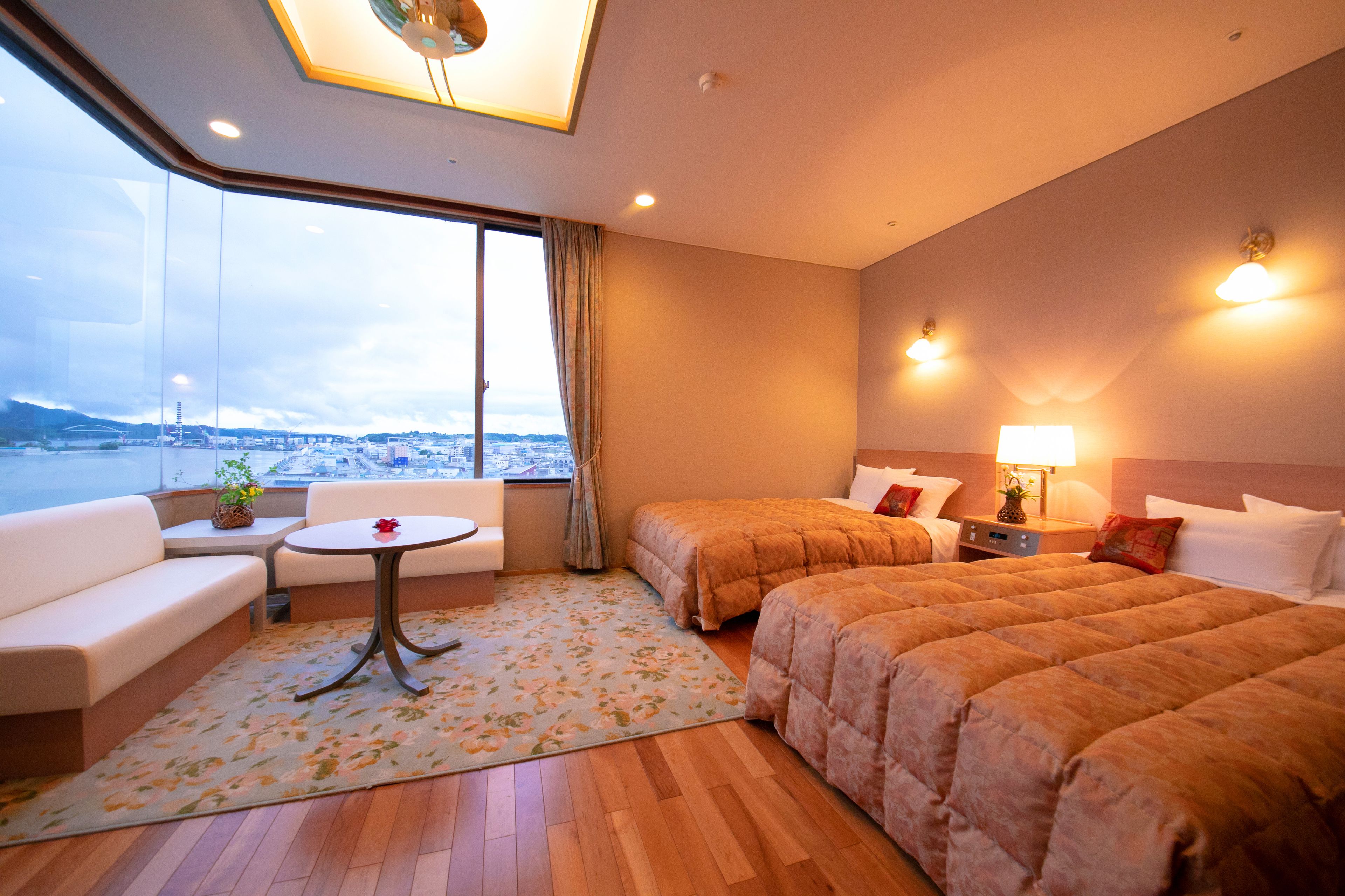 A hotel room with a large window, a sofa, two beds, a small table, and a lamp.