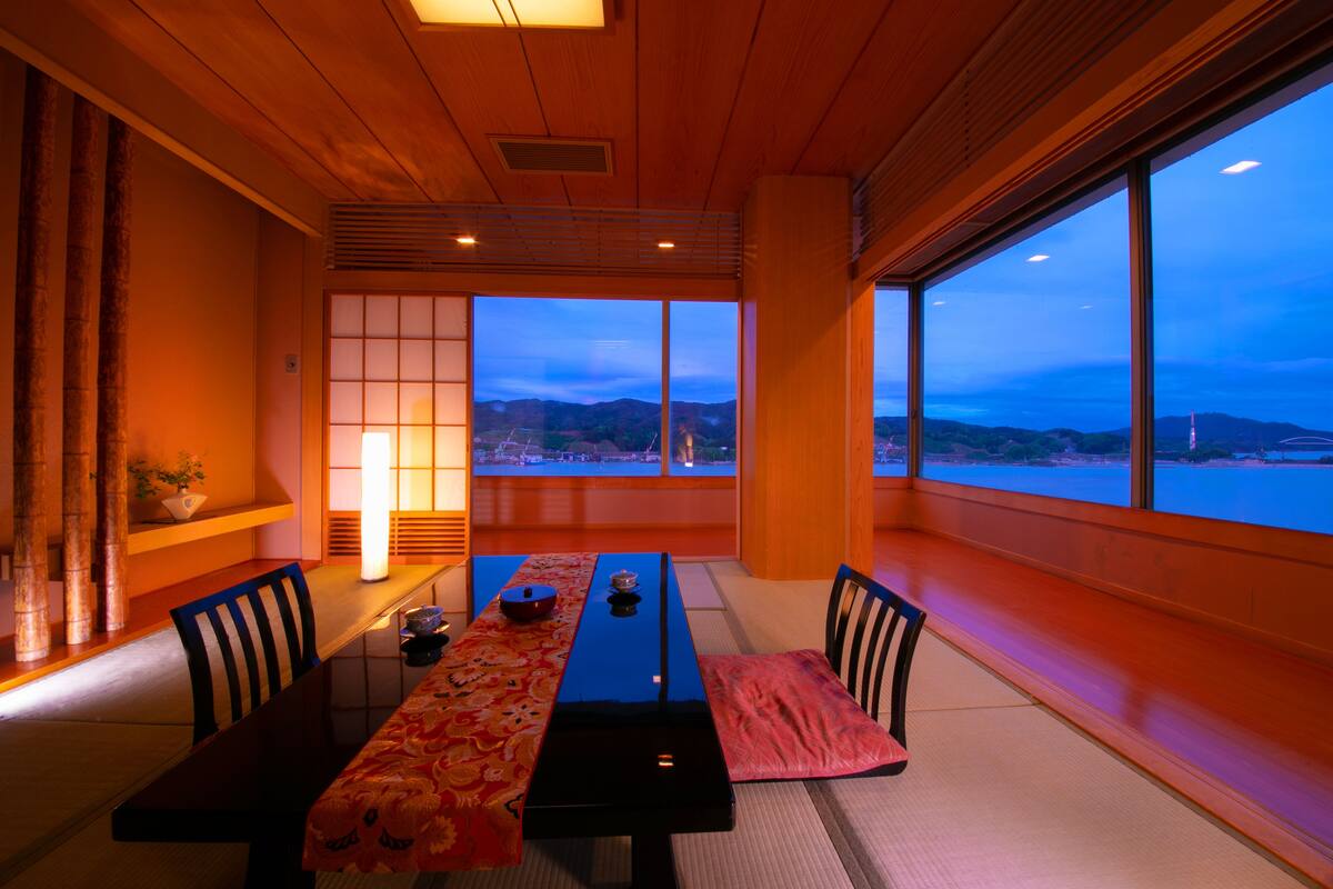 Une pièce de style japonais traditionnel, avec un sol recouvert de tatamis, une table basse agrémentée d’un chemin de table rouge et bleu, et une vue sur la mer et les montagnes, visible à travers de grandes fenêtres.
