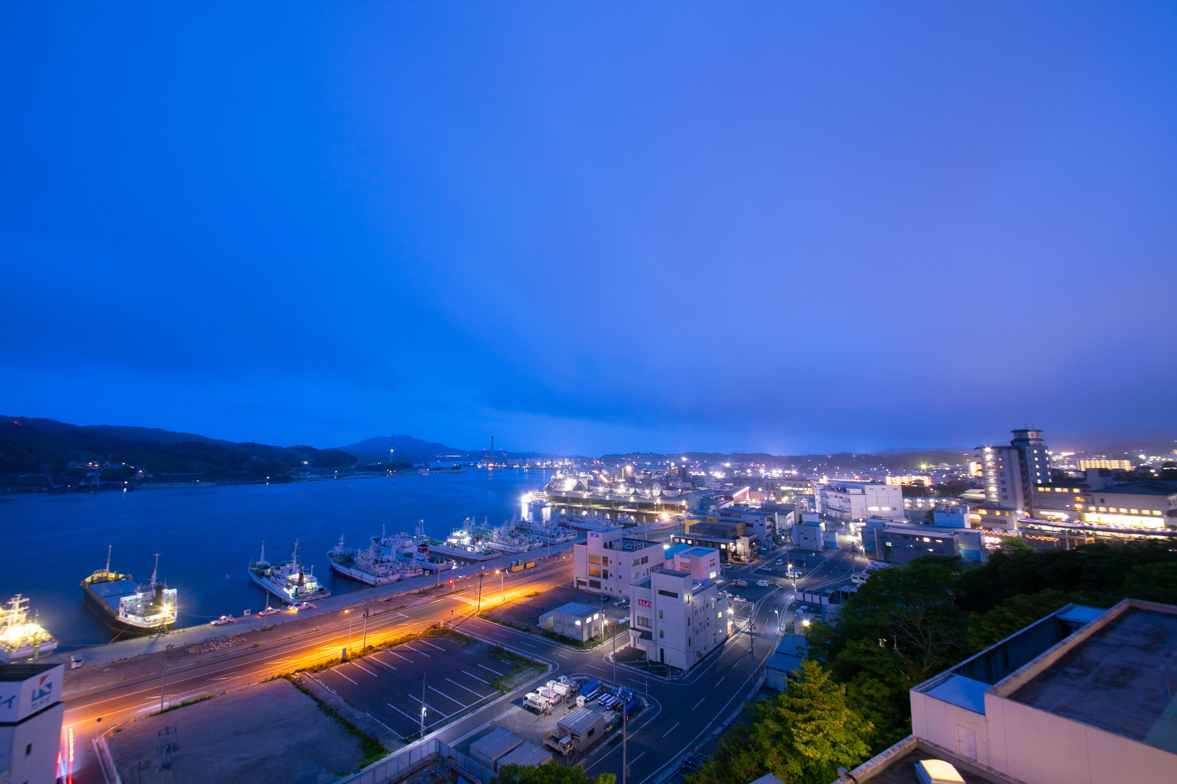 A coastal cityscape at dusk with a marina, industrial buildings, and a highway.