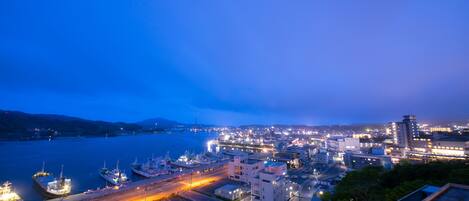 A coastal cityscape at dusk with a marina, industrial buildings, and a highway.
