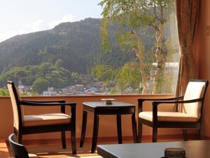 Un balcon avec deux chaises et une petite table, d’où l’on peut admirer un paysage montagneux et contempler une ville située en contrebas.