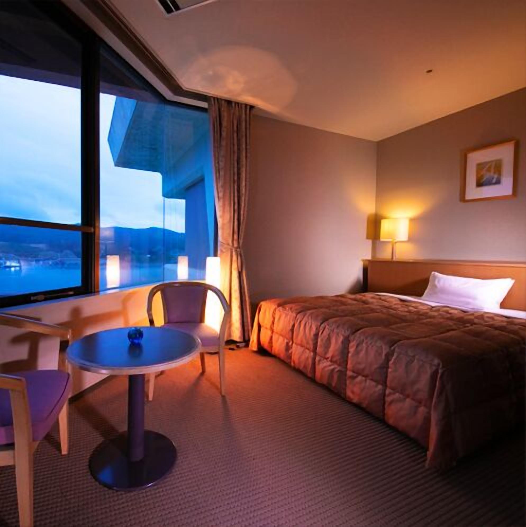 Habitación de hotel con cama, una mesa, dos sillas, ventana con vistas y una lámpara.