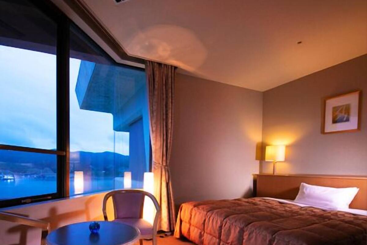Une chambre d’hôtel avec un lit, une table, deux chaises, une fenêtre donnant sur un beau paysage et une lampe.