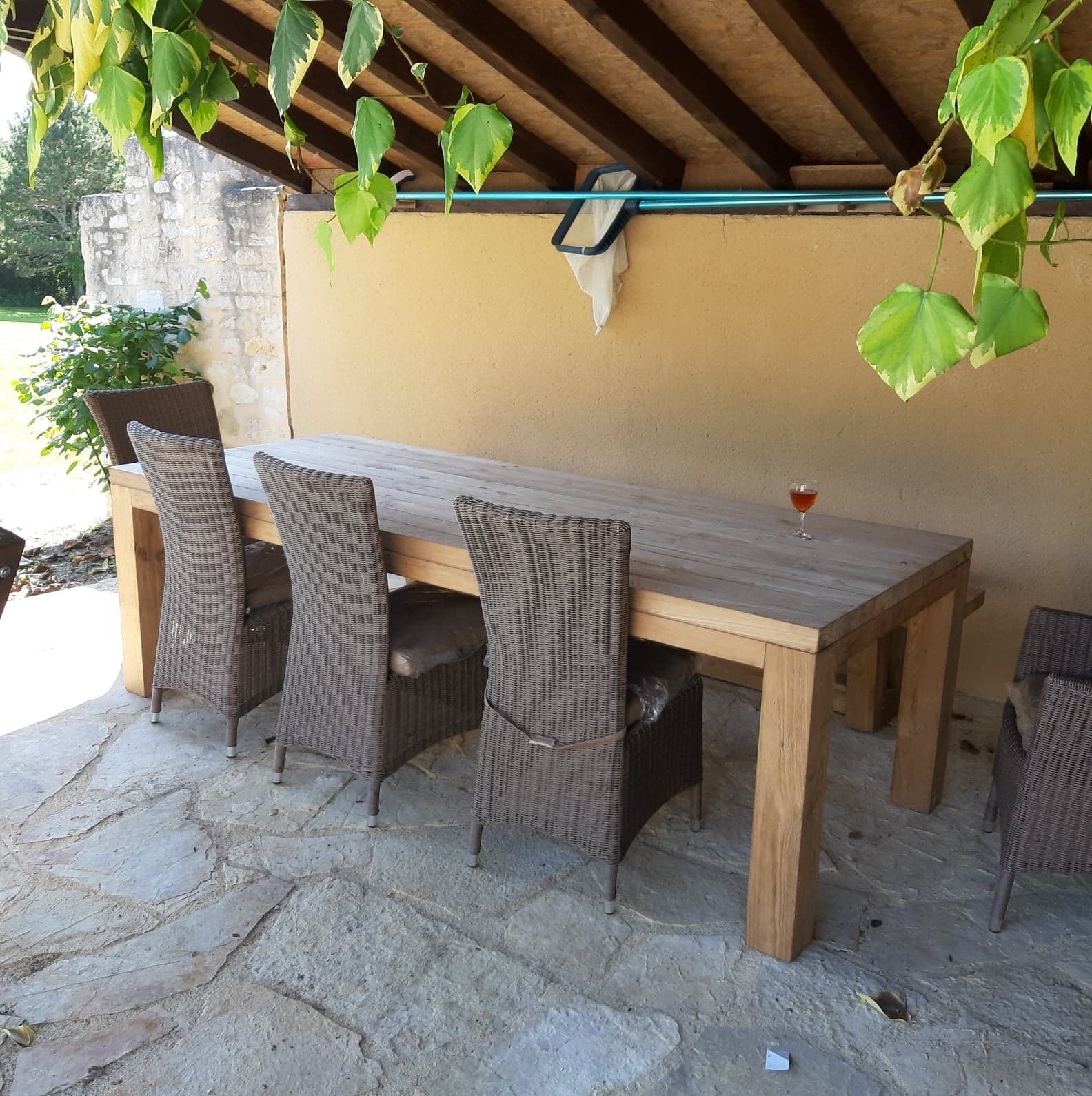Un espace de restauration en plein air avec une table en bois et des chaises en osier, un verre de vin rouge et des plantes suspendues.
