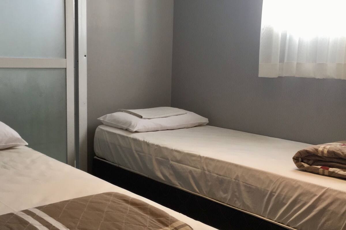 A small, single-room with two beds, one with a quilt and the other with a plain sheet.