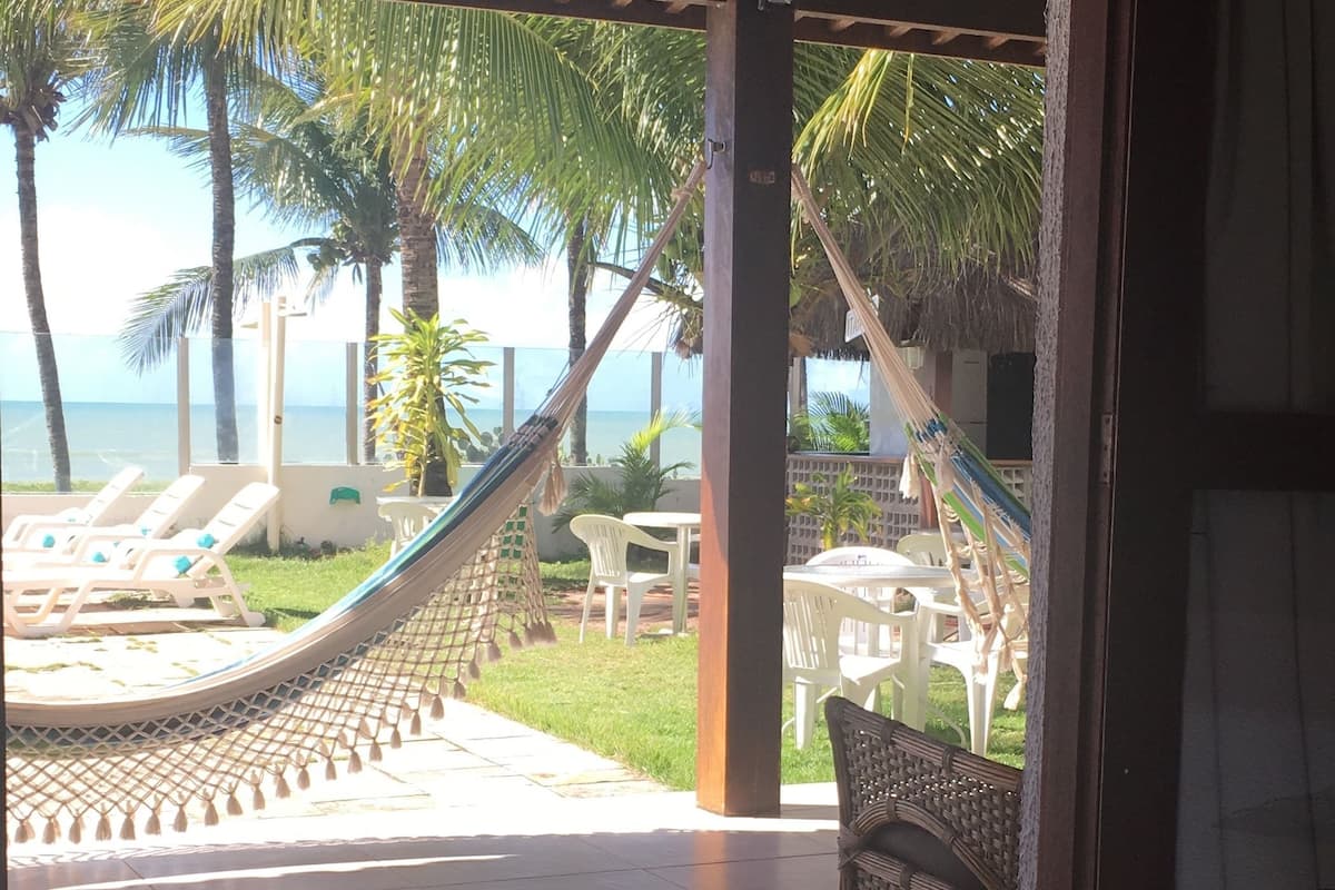 A room with a bed, a view of a beach with palm trees, and a hammock.