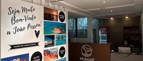 Zona de recepción de un hotel con una pared decorada con fotografías de viajes y un cartel que dice "Seja Muito Bem-Vindô a João Pessoa!"