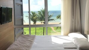 Quarto com uma grande janela com vista para a praia e palmeiras.