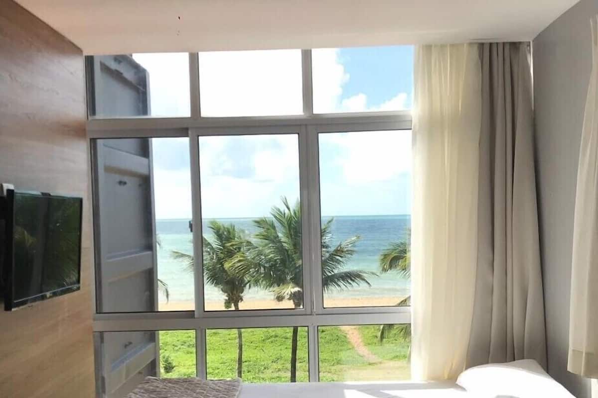 A bedroom with a large window offering a view of the beach and palm trees.