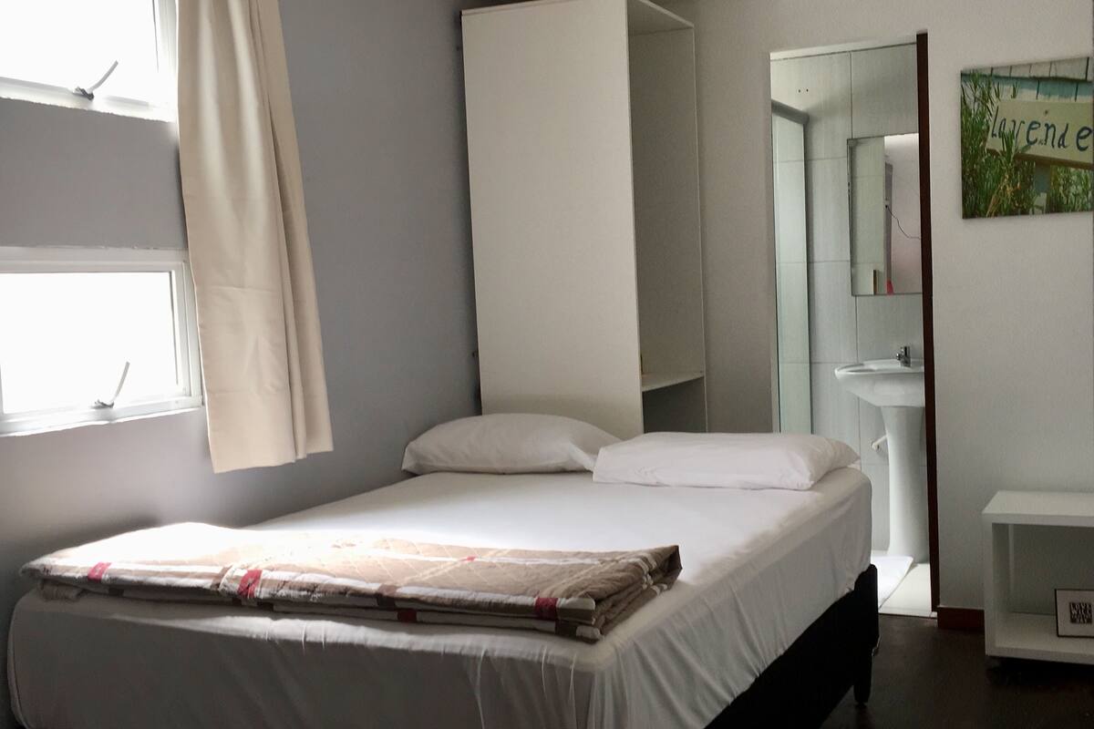 A bedroom with a bed, a window with curtains, a bedside table, a mirror, and a sink.
