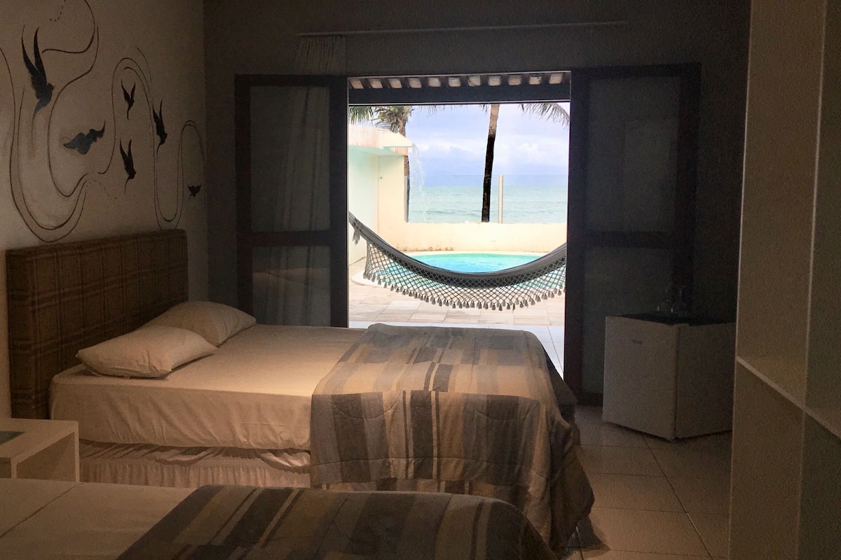 A bedroom with a bed, a nightstand, a view of a beach and pool through an open window.