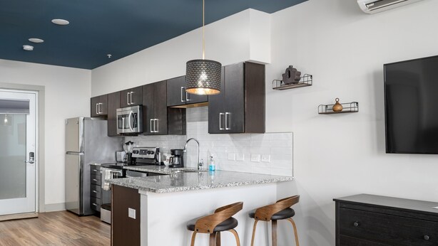 A modern kitchen with dark cabinets, a granite countertop, and two bar stools. - Studio King Suite w/ Kitchen | 103 (Asheville)