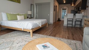 A modern living room with a bed, a round wooden coffee table, and a kitchen area in the background. - Studio King Suite w/ Kitchen | 103 (Asheville)