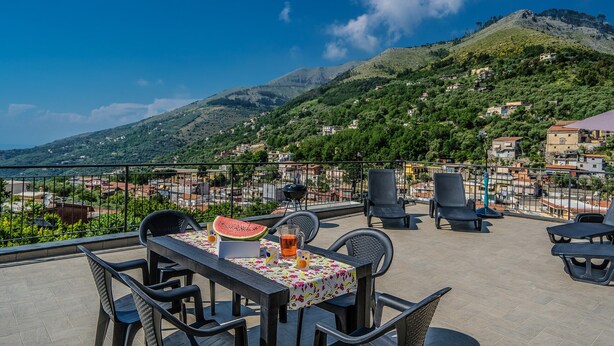 Una terrazza all'aperto con un tavolo apparecchiato per due, sedie e vista su una collina con edifici e uno specchio d'acqua sullo sfondo.