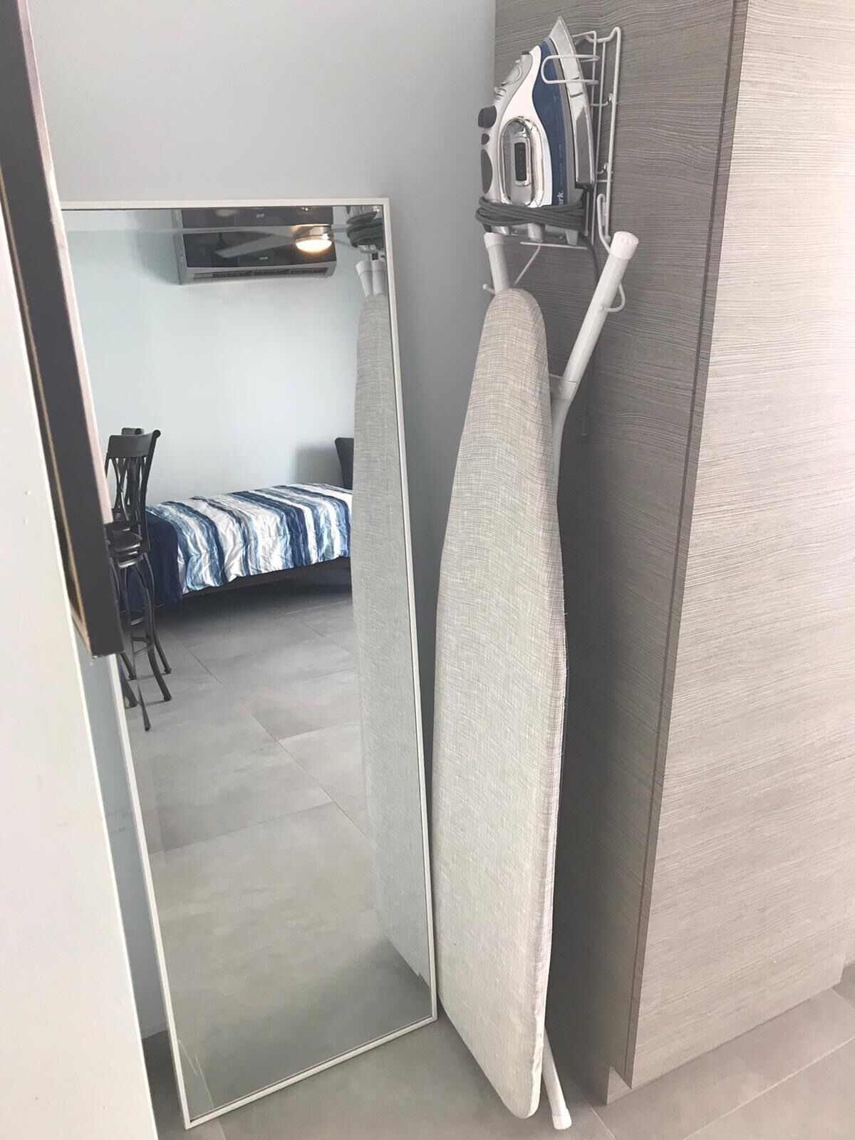 A modern hotel room with a large mirror, an ironing board, and a bed with a blue and white striped bedspread.