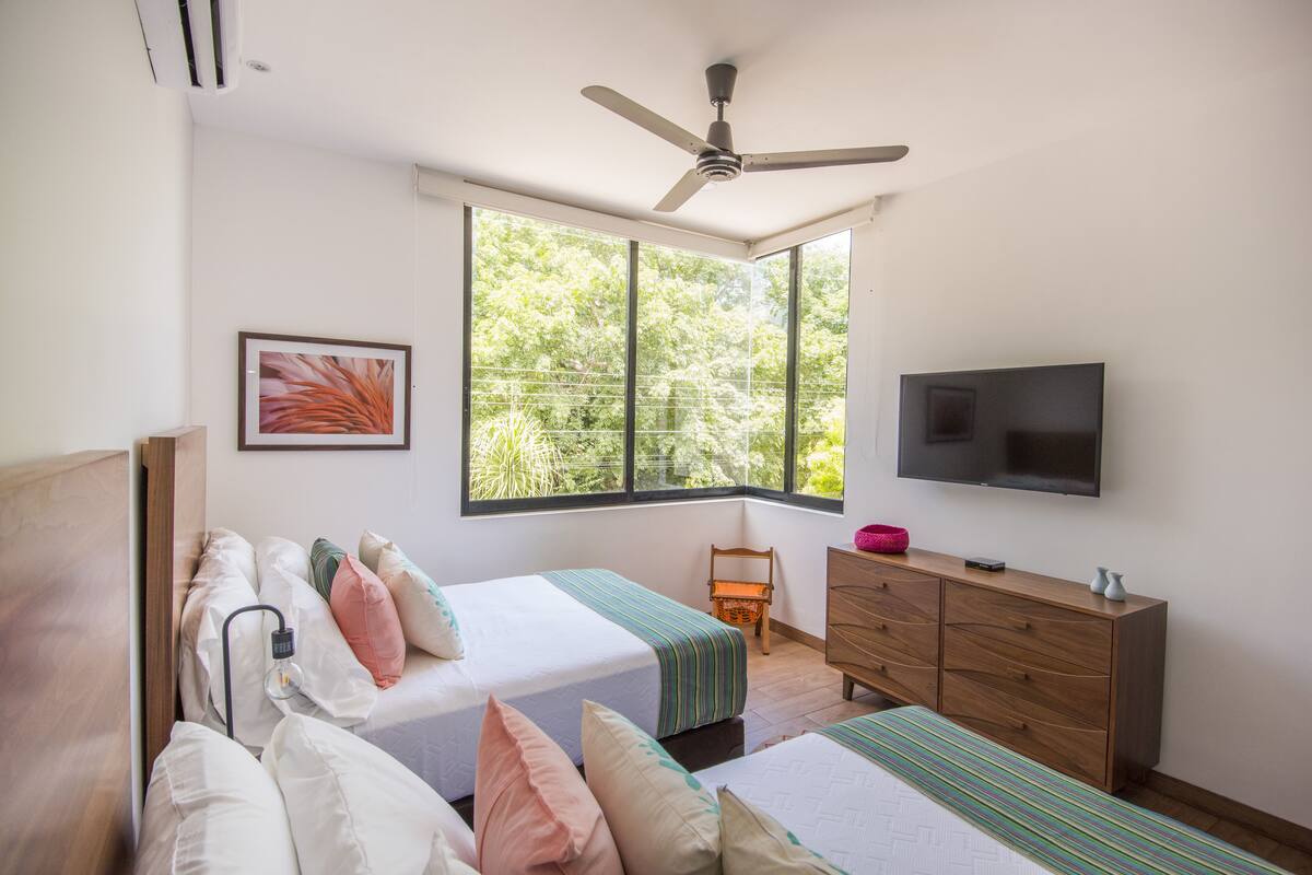 A bedroom with two beds, a TV, a ceiling fan, and a window with a view of trees.