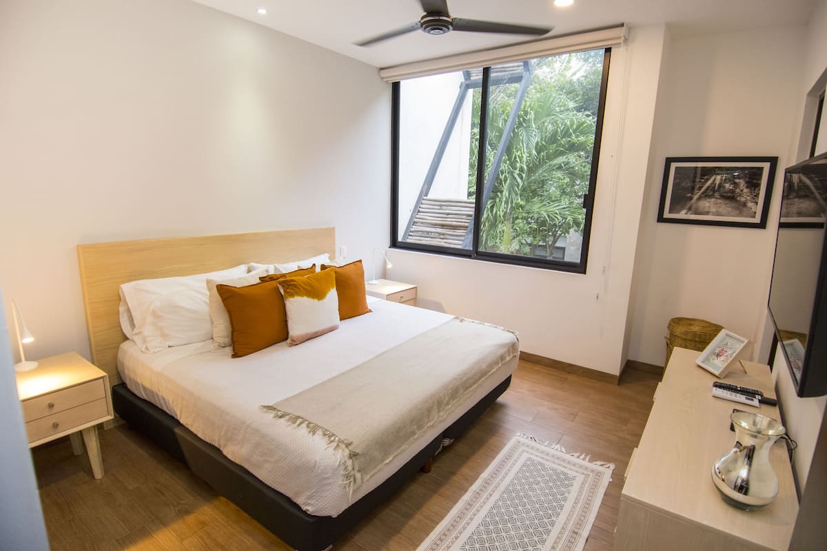 A bedroom with a bed, bedside table, nightstand, window, and a desk with a vase.
