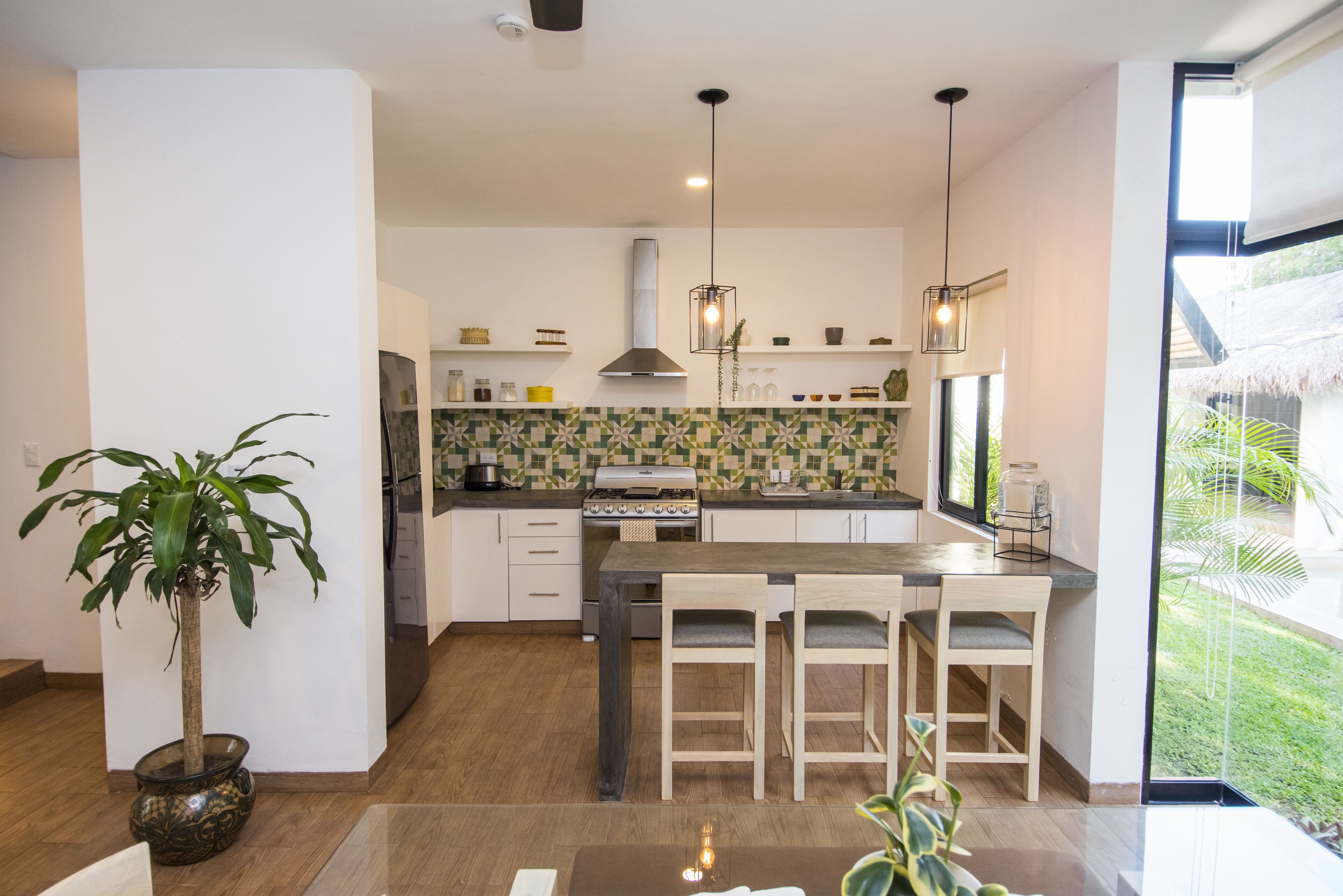A modern kitchen with a central island, wooden chairs, a potted plant, and a view of the outdoors.