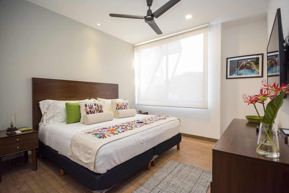A bedroom with a bed, a nightstand, a desk, a chair, a vase with flowers, and a ceiling fan.