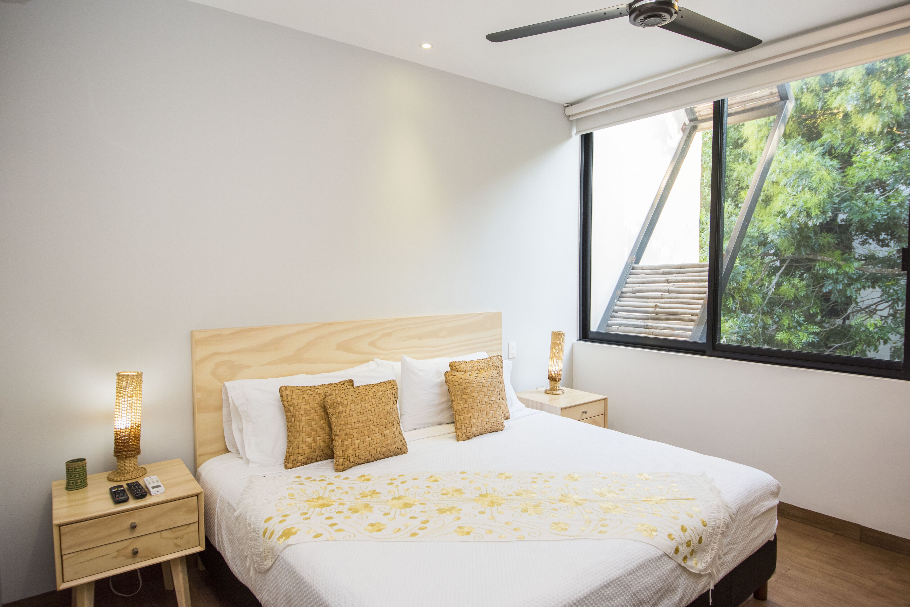 Un lit bien fait, recouvert d’une couvre-lit à motifs floraux blancs et jaunes, deux tables de chevet en bois avec des lampes, et une fenêtre donnant sur un paysage verdoyant.