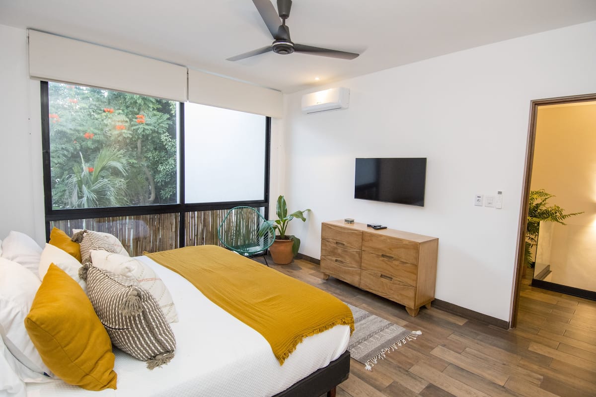 A modern bedroom with a bed, a ceiling fan, a TV, a wooden dresser, and a large window with a view of trees.