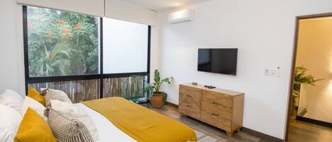 A modern bedroom with a bed, a ceiling fan, a TV, a wooden dresser, and a large window with a view of trees.