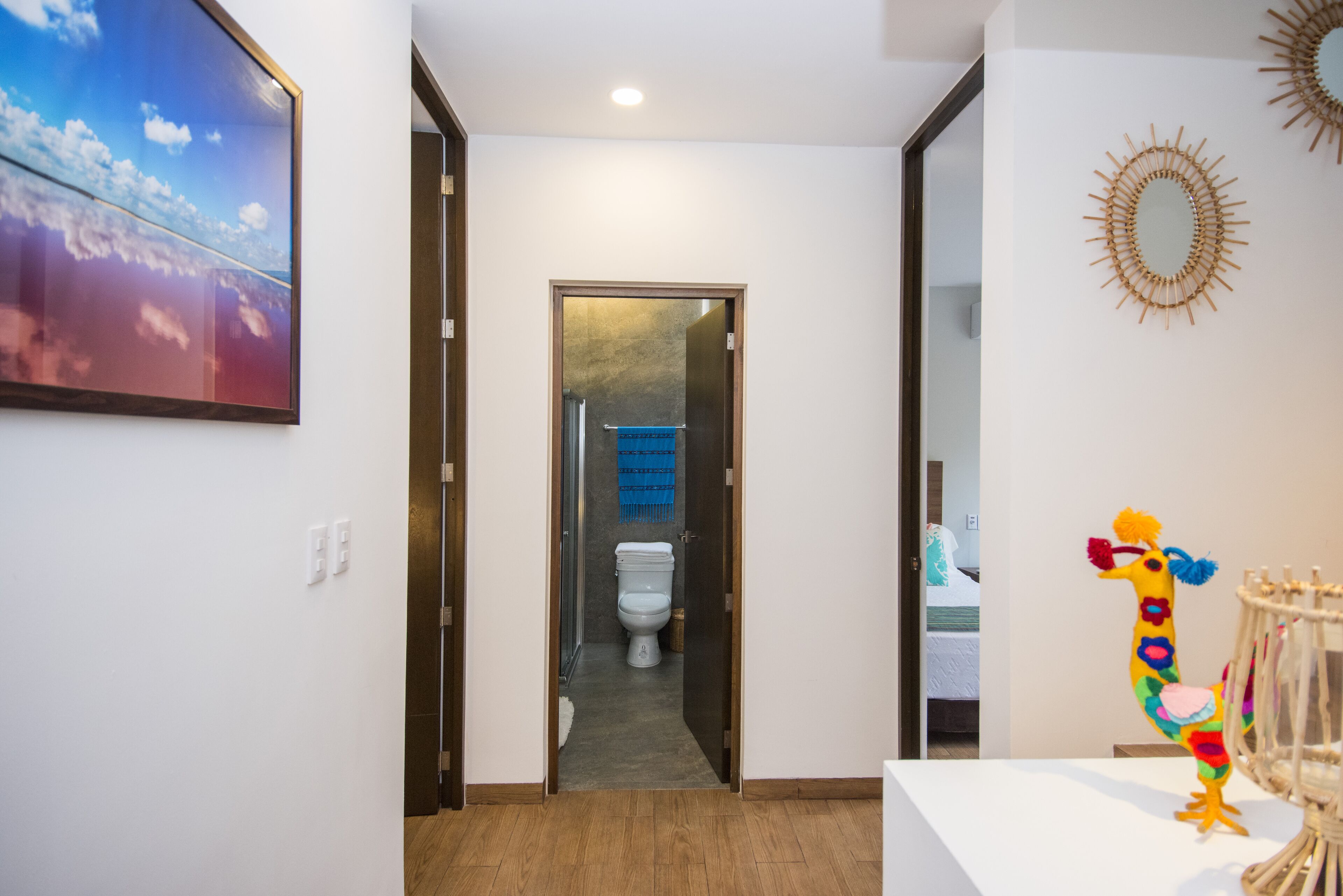 Um corredor com piso de madeira, parede branca e porta que dá para um banheiro com vaso sanitário e toalha azul pendurada.