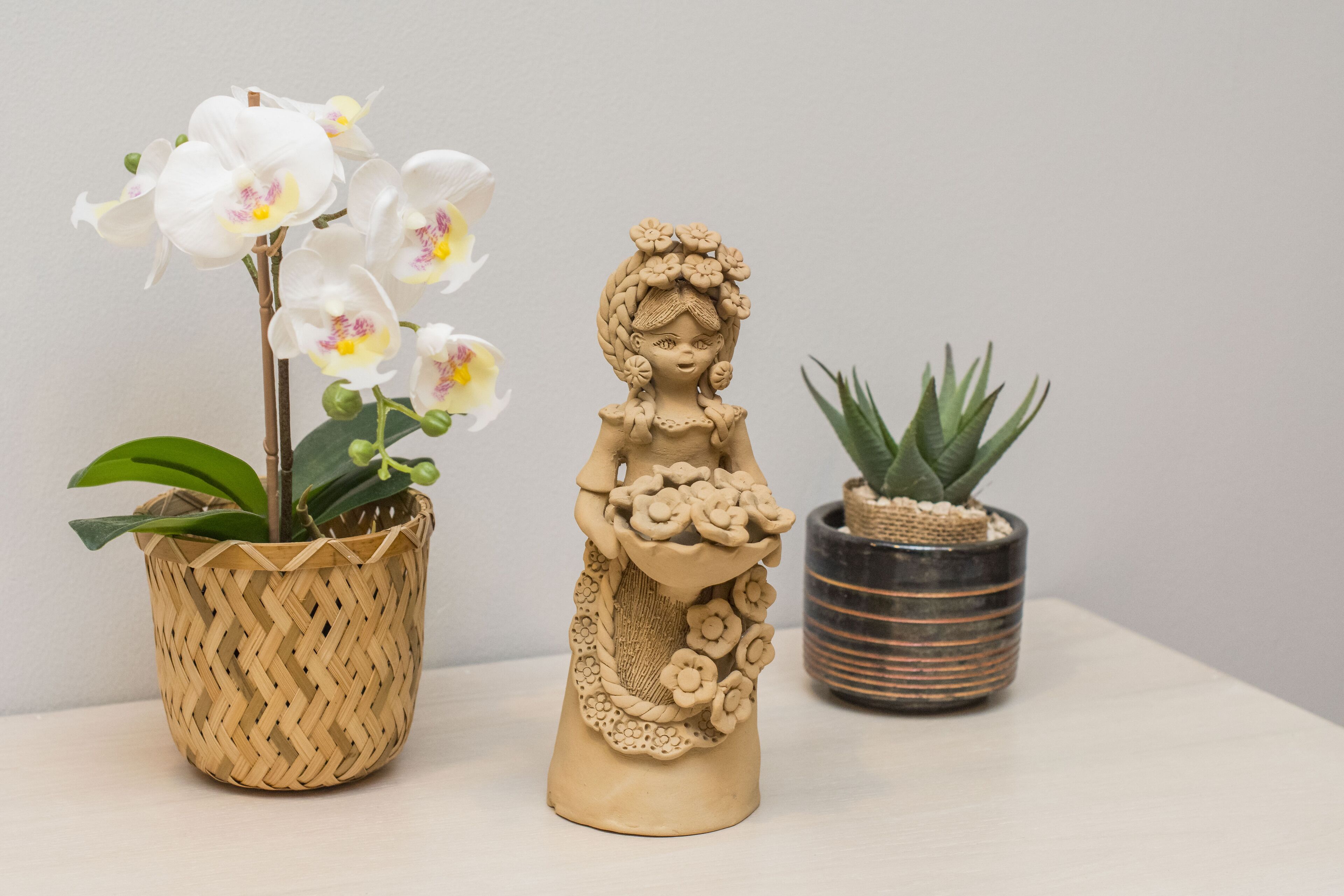 A ceramic figurine of a woman holding flowers, a white orchid plant in a woven basket, and a small cactus plant in a striped pot.