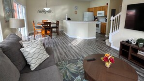 A living room with a gray sofa, a coffee table with a floral rug, a dining area with wooden chairs, a kitchen with white appliances, and a staircase with white railings.