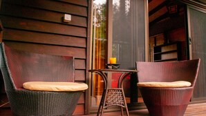 Dos sillas de mimbre con cojines en una terraza de madera, una mesita con una vela y un ventanal corredero que da a un espacio interior.