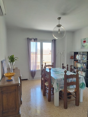 A dining area with a table, chairs, a bookshelf, and a view through a window.