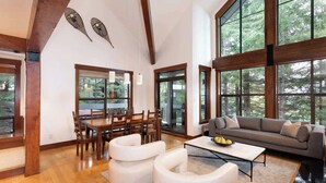 Ein geräumiges Wohnzimmer mit einem Holzesstisch, weißen Sofas und großen Fenstern mit Blick auf den Wald.