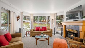 A modern living room with a sofa, armchairs, a fireplace, and large windows overlooking a garden. - Forest condo w/ AC, pool & gym (Whistler)